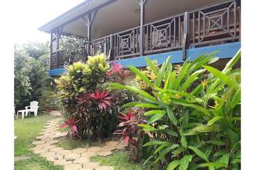 A tropical garden surrounds a two-story house with a wooden balcony. Colorful plants and greenery line a stone pathway. White chairs sit in the yard.