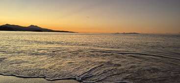 Golden sunset over a calm sea, gentle waves lapping at the shore. Silhouetted mountains in the distance, peaceful, serene atmosphere. Warm, pastel colors fill the sky.