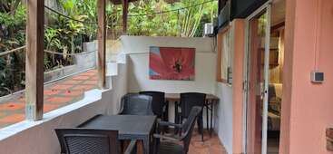 This image shows a cozy outdoor patio with black chairs and tables, surrounded by lush greenery. A floral painting decorates the wall. Glass door entrance visible.