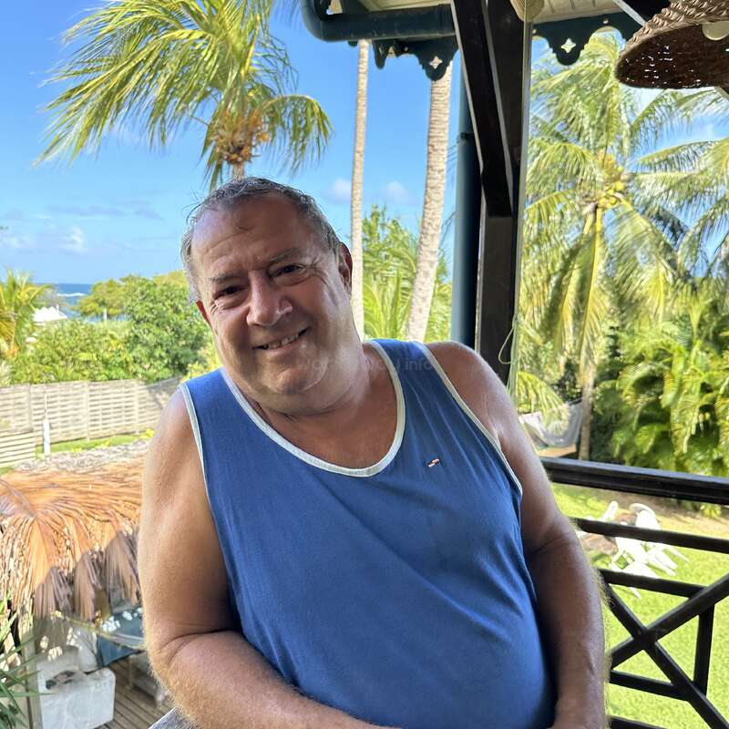 Um homem sorridente, vestindo uma camiseta regata azul, está em uma varanda, cercado por palmeiras verdejantes e uma paisagem tropical, sob um céu azul brilhante.