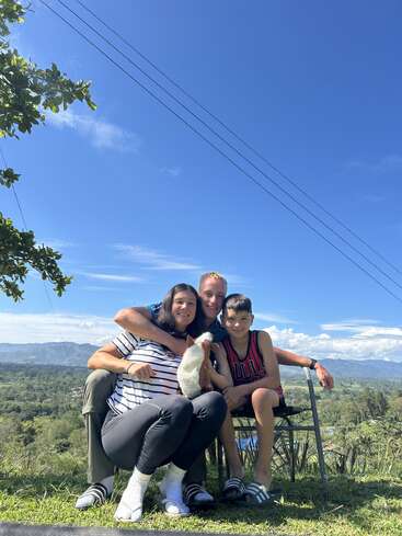 A smiling family of three sits outdoors on a sunny day, with a picturesque mountain landscape and clear blue sky in the background. They look happy.