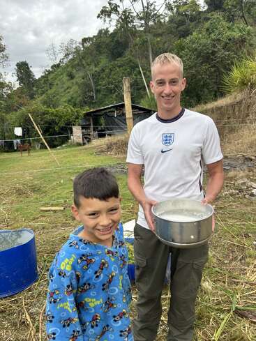 A smiling man in an England shirt holds a pot of liquid, standing outdoors with a cheerful boy in blue pajamas, surrounded by lush greenery and hills.