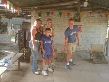 Four people pose and smile in a rustic, open space with festive bunting. Two cornhole boards sit behind them. It's sunny, relaxed, and casual.