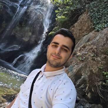 Un homme prend un selfie devant une magnifique cascade, au milieu des rochers et de la verdure, souriant et profitant d'un moment de tranquillité en pleine nature.
