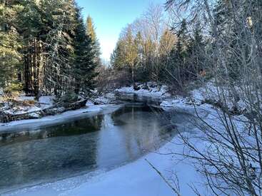 Eine friedliche Winterszene mit einem teilweise zugefrorenen Fluss, der sich durch einen verschneiten Wald schlängelt. Hohe immergrüne Bäume säumen das Ufer und spiegeln das Sonnenlicht unter einem strahlend blauen Himmel wider.