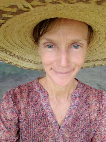 Eine Frau lächelt warm in die Kamera. Sie trägt einen breitkrempigen Strohhut und eine blumengemusterte Bluse im Vintage-Stil und steht an einem leicht bewölkten Tag im Freien.