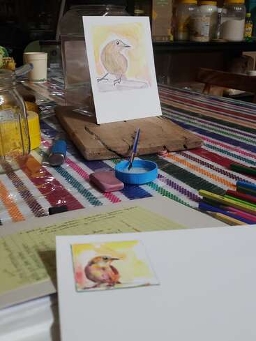 Auf einem Tisch mit Malutensilien und bunten Stiften liegt ein Aquarellbild von einem Vogel auf einem Holzbrett, daneben das Referenzfoto.