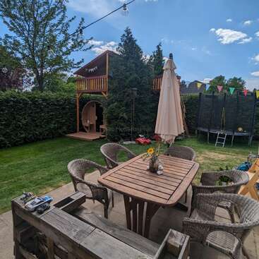 The image depicts a backyard with a patio table and chairs, a playhouse, a trampoline, and a hedge, set against a blue sky with white clouds.