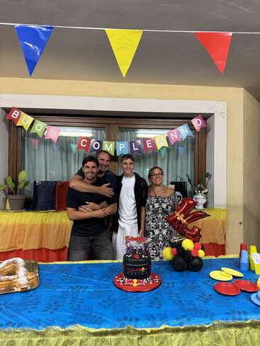 Quatro pessoas posam felizes em frente a um banner "Buon Compleanno", cercadas por decorações coloridas, um bolo da Red Bull Racing, doces e balões festivos em uma festa de aniversário.