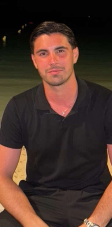 Um homem de cabelos escuros com uma camisa polo preta está sentado em uma praia à noite, sorrindo suavemente. Luzes fracas refletem na água atrás dele.