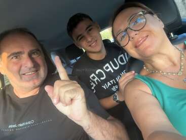 Um grupo sorridente de três pessoas, dois adultos e um adolescente, sentados juntos dentro de um carro. O homem gesticula com um dedo, todos parecem felizes.