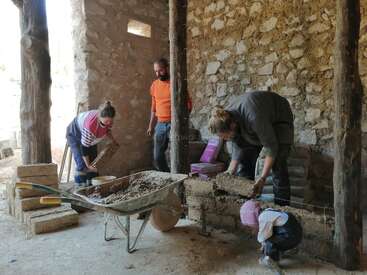 Três adultos e uma criança estão construindo com materiais naturais dentro de uma estrutura de pedra rústica, usando terra, vigas de madeira e tijolos de sabugo, com um carrinho de mão por perto.