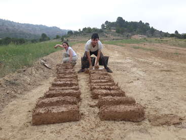 Duas pessoas estão ao ar livre, fazendo alegremente tijolos de adobe com lama. Elas estão cercadas por campos e colinas, com fileiras de tijolos recém-formados secando no chão.