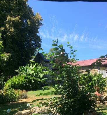 The image depicts a serene stone house with a red roof, nestled among lush greenery and trees, set against a clear blue sky on a sunny day.