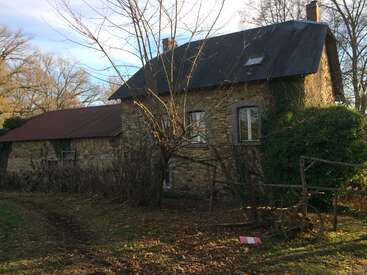 The image depicts a rustic stone house with a dark roof, surrounded by trees and bushes, featuring a chimney and a small window on the upper floor.