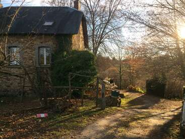The image depicts a charming stone house with a dark roof, nestled among trees and bushes, featuring a dirt path leading to the entrance.