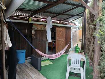 Esta imagen muestra una acogedora zona exterior con una hamaca rosa, sillas de plástico, una mesita, una alfombra verde, un cobertizo de madera y varios objetos personales que crean un ambiente relajado.