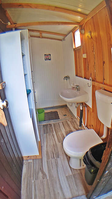 Esta imagen muestra un pequeño cuarto de baño rústico con paredes y techo de madera, inodoro blanco, lavabo, estantería, papelera y cubo verde sobre suelo de baldosas. Iluminación brillante.