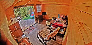 Esta acogedora cabaña de madera cuenta con una cama individual con ropa de cama de colores, un escritorio, una silla y una puerta abierta que da a una terraza soleada y al jardín exterior.