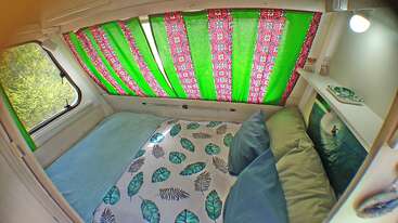 El acogedor interior de la caravana cuenta con una cama perfectamente hecha con ropa de cama estampada con hojas, cortinas verdes y rosas, almohadas mullidas, estanterías con libros y una foto de un surfista.