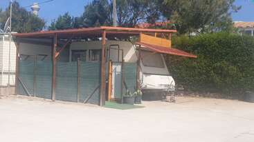 Una caravana aparcada se encuentra bajo un cobertizo de madera con un toldo que da sombra. La rodean vallas verdes y plantas en macetas. Los árboles y los setos proporcionan privacidad y sombra.