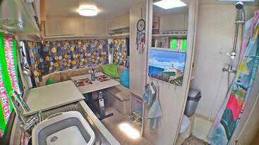 El acogedor interior de esta caravana cuenta con una cocina compacta, cortinas de colores, un comedor con cojines, un atrapasueños, cuadros decorativos, un pequeño cuarto de baño con ducha y toques decorativos vibrantes por todas partes.