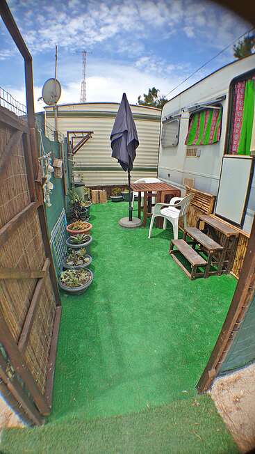 Una acogedora zona exterior con césped artificial, plantas en macetas, una mesa de madera, sillas de plástico blancas, escalones de madera, una sombrilla y ventanas decoradas en verde junto a una caravana.