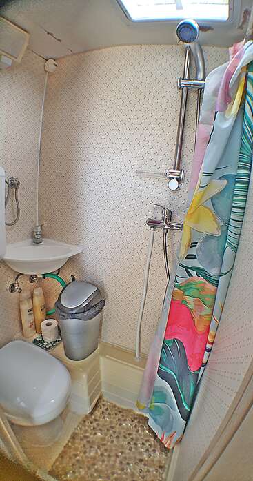 Se trata de un cuarto de baño compacto con ducha, cortina de ducha de colores, lavabo pequeño, inodoro, papelera y artículos de aseo, probablemente en una casa móvil o un barco.