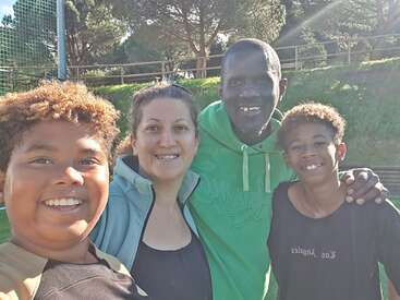 Cuatro personas sonrientes, probablemente una familia, posan al aire libre en un día soleado. Están de pie muy juntas, con árboles verdes, césped y una valla de madera al fondo.