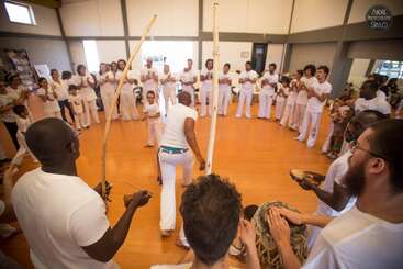 A group of people in white clothes form a circle, playing instruments and clapping, while two individuals perform capoeira moves in the center on a wooden floor.