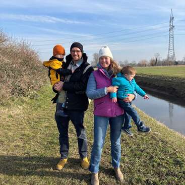 The image depicts a family of four standing on a grassy bank beside a river, with the man holding a child in his arms and the woman holding another child.