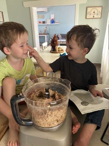 Two young boys sit on a kitchen counter, happily baking together. One boy playfully touches the other's nose, next to a food processor with dough.