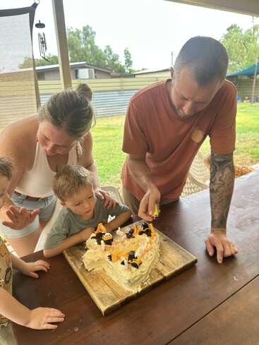 A family gathers around a heart-shaped birthday cake decorated with toy diggers. An adult lights candles while two young boys watch eagerly, excited for the celebration.