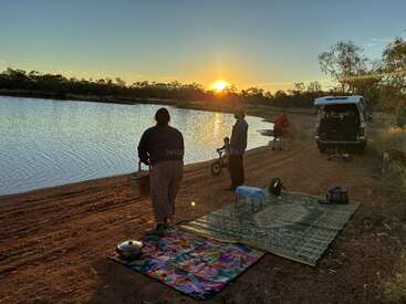 Al atardecer, a orillas de un lago, una familia disfruta de un picnic. Las esteras, la cesta, la furgoneta y las bicicletas crean un ambiente relajado al aire libre en plena naturaleza.