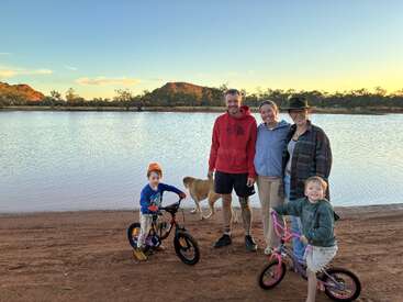 Una familia de cinco miembros, entre los que se encuentran dos niños con bicicletas y un perro, posa junto a un pintoresco lago al atardecer, con colinas y árboles al fondo.