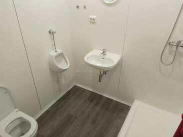 This image shows a clean, minimalistic bathroom with a toilet, urinal, wall-mounted sink, shower area, white walls, and dark wood-patterned flooring, all neatly arranged.