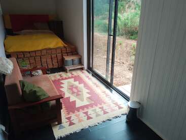 A cozy room with a bed covered in a yellow blanket, a patterned rug, a small sofa, sliding glass doors, and stacks of bricks underneath the bed.