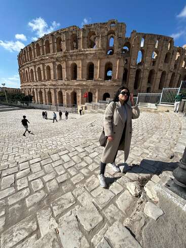 Una mujer con abrigo beige posa con confianza delante del antiguo anfiteatro romano de El Djem, Túnez, bajo un cielo azul brillante.