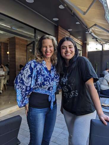 Dos mujeres posan muy juntas, sonrientes, a la salida de un café. Una lleva una blusa azul de flores y vaqueros, la otra una camiseta negra y pantalones claros. Ambiente informal y alegre.