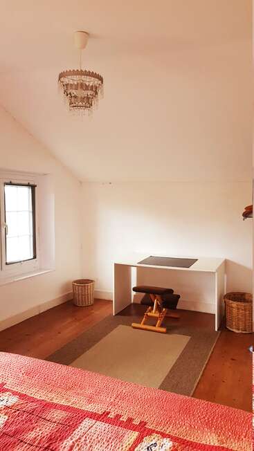 This image shows a cozy, minimalist room with white walls, wooden floor, chandelier, desk, ergonomic stool, two woven baskets, window, and a red bedspread.