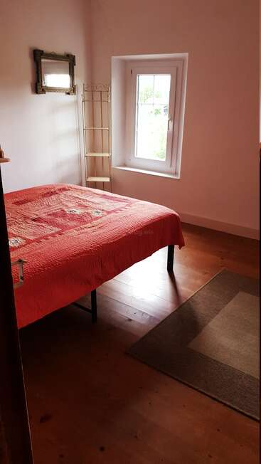 A cozy bedroom with wooden floor, a red bedspread, small window, ornate mirror, simple shelf, white walls, and a brown carpet creating a warm atmosphere.