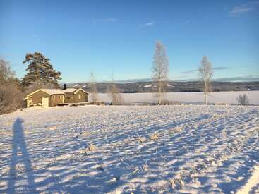 Ein gemütliches Holzhaus steht inmitten eines verschneiten Feldes unter strahlend blauem Himmel. Kahle Bäume stehen hoch, und ein langer Schatten erstreckt sich über die Landschaft.