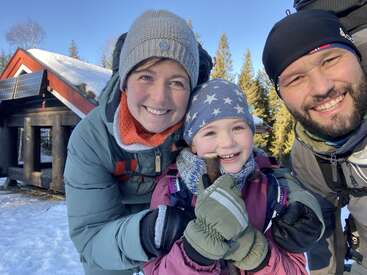 The image shows a family of three smiling at the camera, with a woman, a child, and a man, all wearing winter clothing, posing together outdoors.