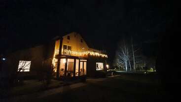 Una acogedora casa brilla cálidamente por la noche, adornada con guirnaldas de luces a lo largo del balcón. Las ventanas iluminadas revelan interiores acogedores, mientras que el patio circundante está oscuro y tranquilo.