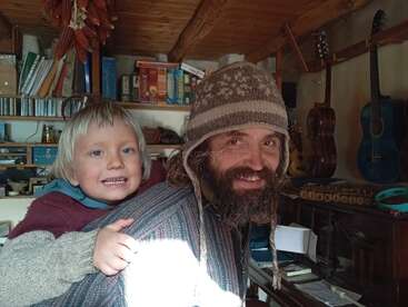 Um cara sorridente usando um gorro de tricô e uma criança de cabelo claro estão dentro de casa, rodeados por livros, violões e móveis de madeira aconchegantes, banhados pela luz do sol.