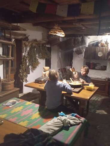Duas crianças sentadas em uma mesa rústica de madeira, compartilhando uma refeição em uma cozinha aconchegante e mal iluminada, com ervas penduradas, cestas de vime e tecidos coloridos ao redor.