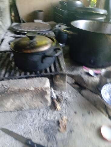 Essa imagem mostra uma cena rústica de cozinha com panelas e frigideiras grandes em um fogão de pedra ou concreto, cinzas espalhadas e alguns utensílios de cozinha por perto.