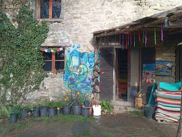 Uma casa rústica de pedra com hera, murais coloridos nas paredes, vasos de plantas, bandeiras de oração e fitas penduradas. Arte, natureza e toques vibrantes criam uma atmosfera aconchegante e artística.