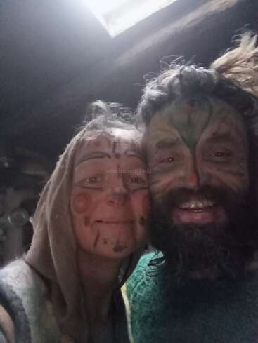 Um homem e uma mulher sorridentes, com pinturas faciais coloridas, tiram uma selfie em close-up. A mulher usa um capuz, criando uma atmosfera divertida e alegre.