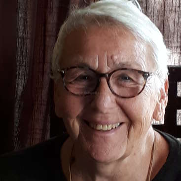 L'image représente une femme âgée aux cheveux blancs courts, portant des lunettes à monture foncée et une chemise noire, sur fond de rideau marron.